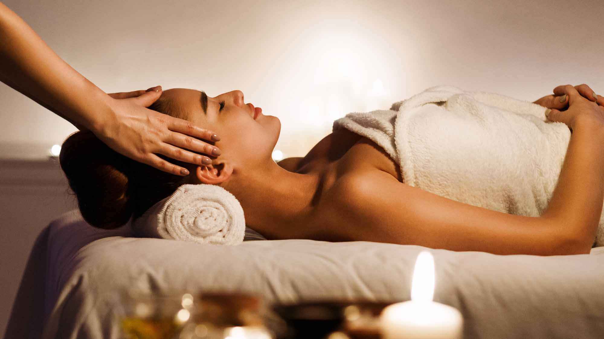 Young woman enjoying face massage with aromatic candles in spa salon